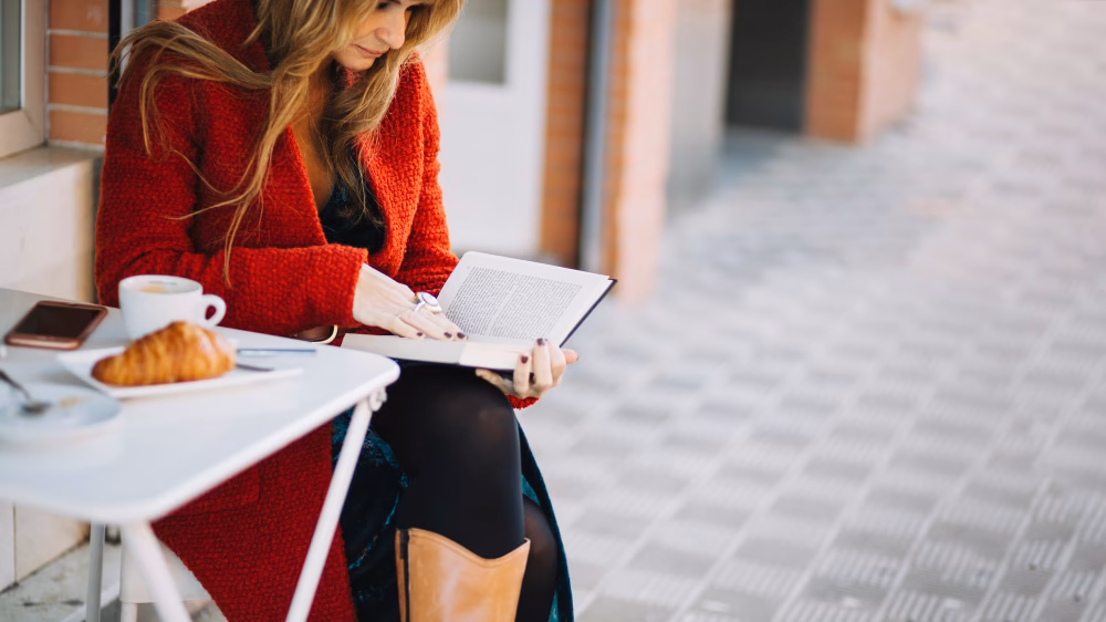 Catherine m. Szexuális élete könyvismertetőhöz képi illusztráció. Egy elegánsan öltözött nő olvas egy kávézó asztalánál.