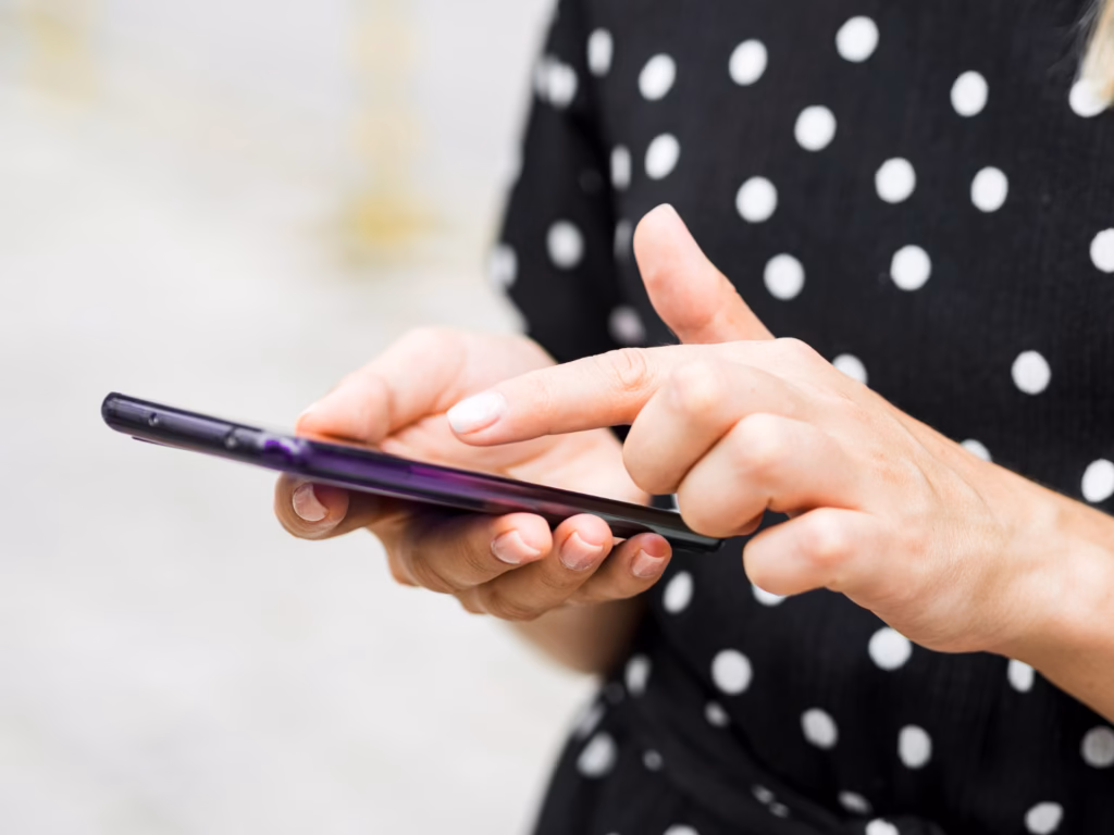 a fotón egy női kéz látható, amint éppen egy mobil telefonon navigál. A fotó illusztráció a távirányítós vibrátorokról szóló blog bejegyzéshez.