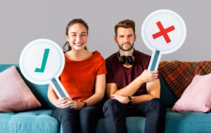 Igen-nem lista készítése pároknál. A képen egy férfi és egy nő ül egy sofán, a férfi kezében egy nemet jelző tábla van, a nő kezében pedig egy igent jelző, pipa tábla van.